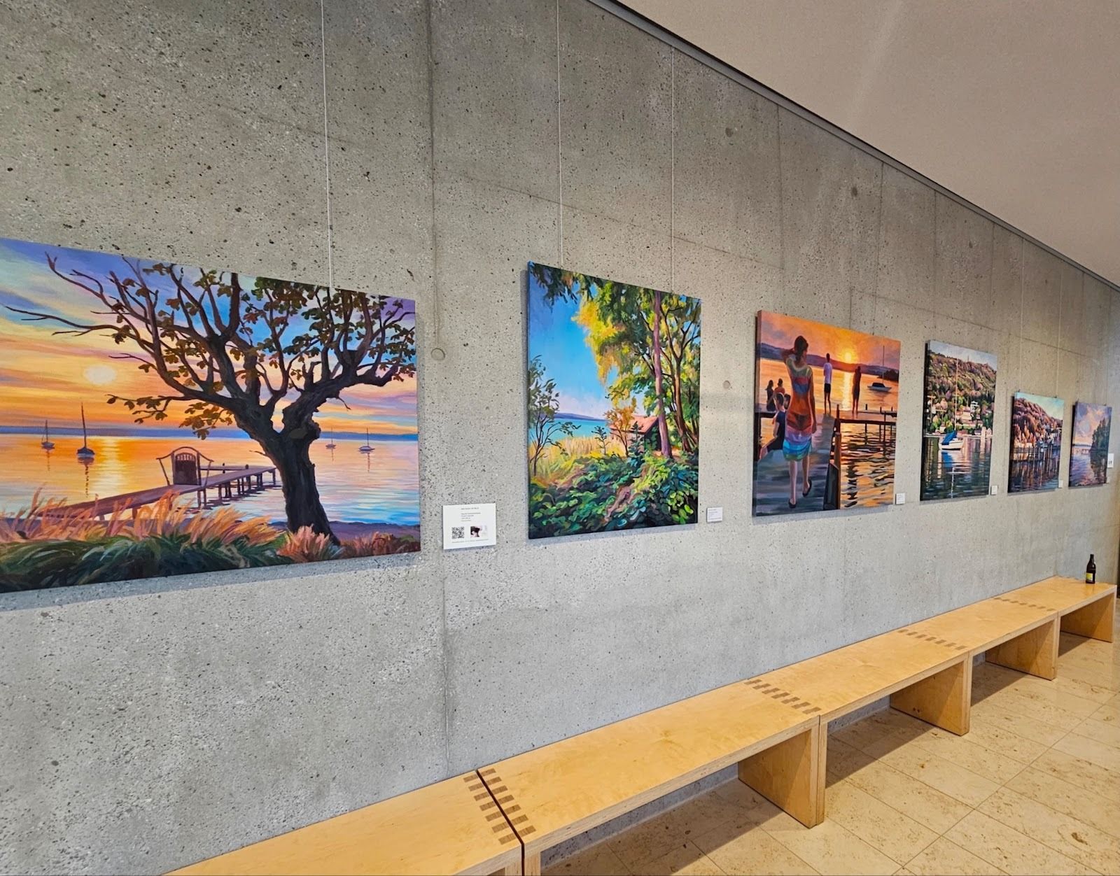 Eine Galeriewand mit fünf Landschaftsgemälden, die über einer langen Holzbank hängen.