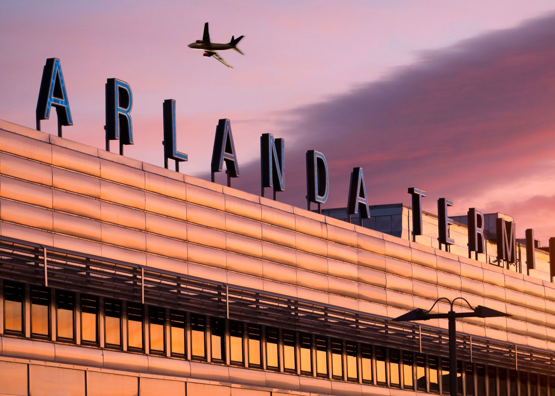 Arlanda Airport building at sunset, plane flying overhead.