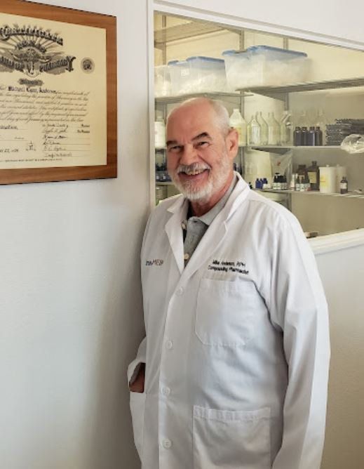 Man in lab coat smiles, stands by a wall with a framed certificate. Laboratory interior in background.