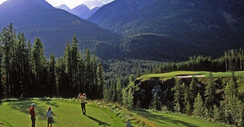 Golfer teeing off on a lush green course with mountain peaks in the background.
