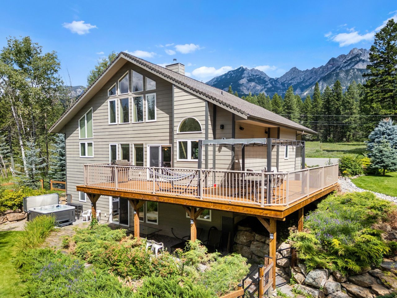 Two-story home with a large wooden deck, stone foundation, and hot tub, set against a backdrop of mountains and forest.