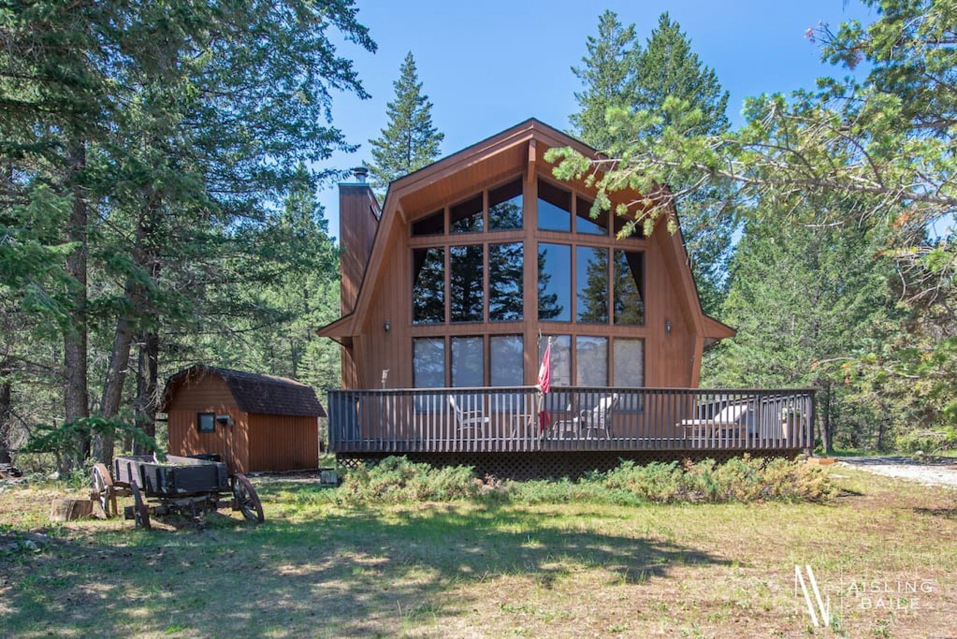 Brown wooden A-frame cabin with large windows and a wraparound deck in a wooded area.