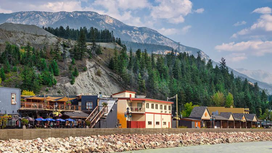 A small town with buildings nestled at the base of a forested mountain, fronted by a rocky shoreline and calm water.