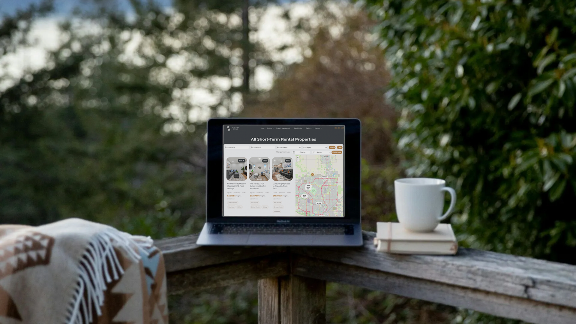 A laptop displaying a map and property listings sits on a wooden deck next to a coffee mug and a patterned blanket.