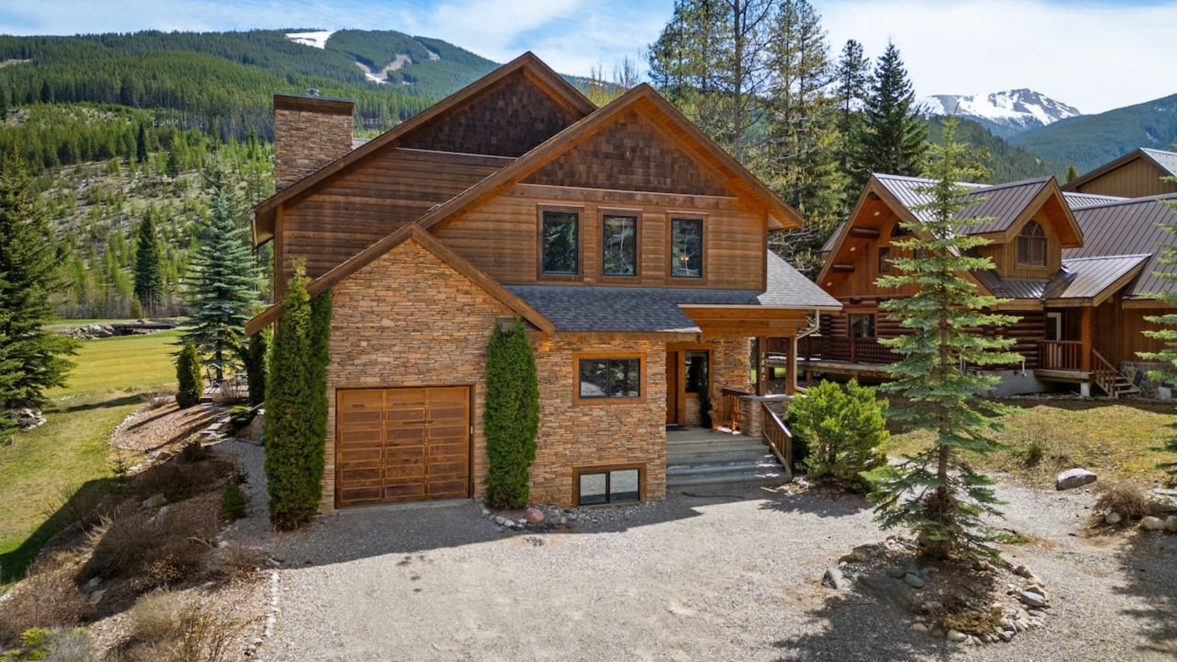 A two-story mountain home with wood and stone siding, a garage, and a gravel driveway set against a forested mountain.