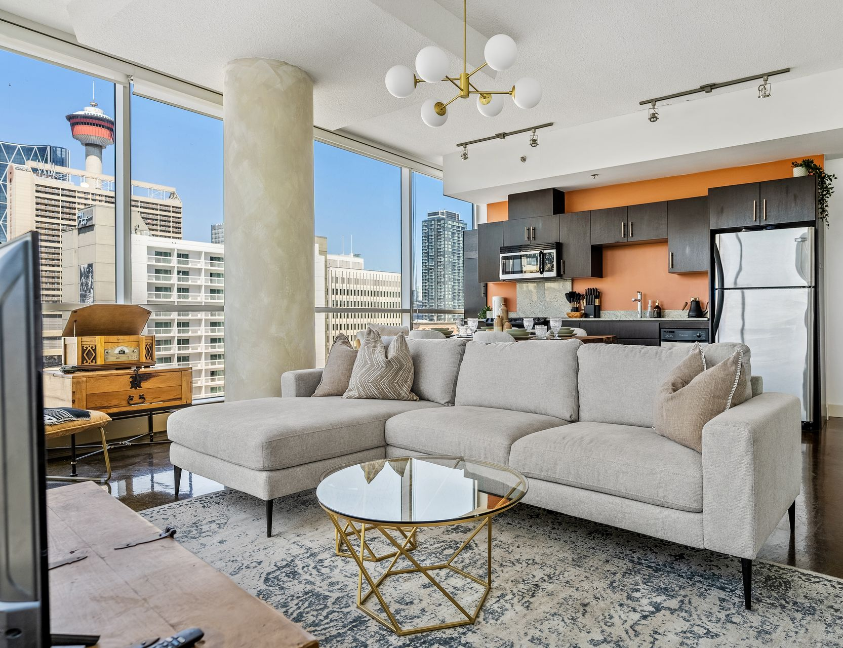 Bright living room with city view, beige sectional sofa, glass coffee table, and kitchen in the background.