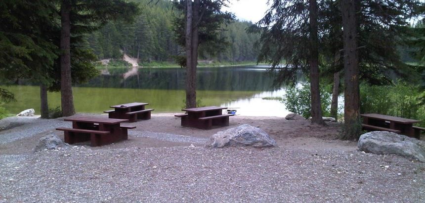 Picnic tables by a lake in a wooded area, with gravel ground and trees surrounding.