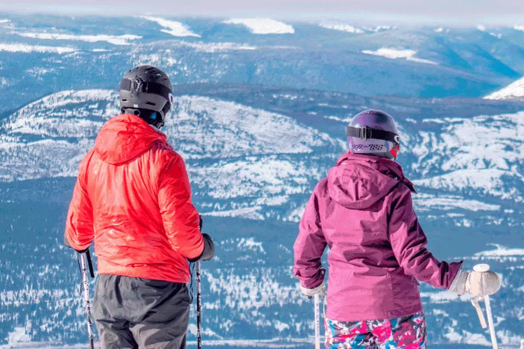 Two people in bright jackets look over a snowy mountain landscape from a ski slope