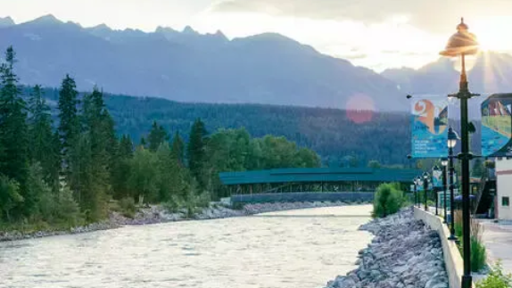 A river flows toward a covered bridge with mountains in the background under a sunset sky, with street lamps in the front.