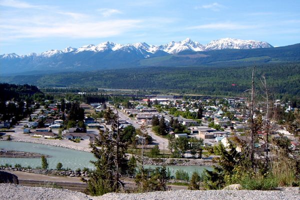 A small town nestled in a valley beside a turquoise river, with snow-capped mountains under a blue sky in the background.