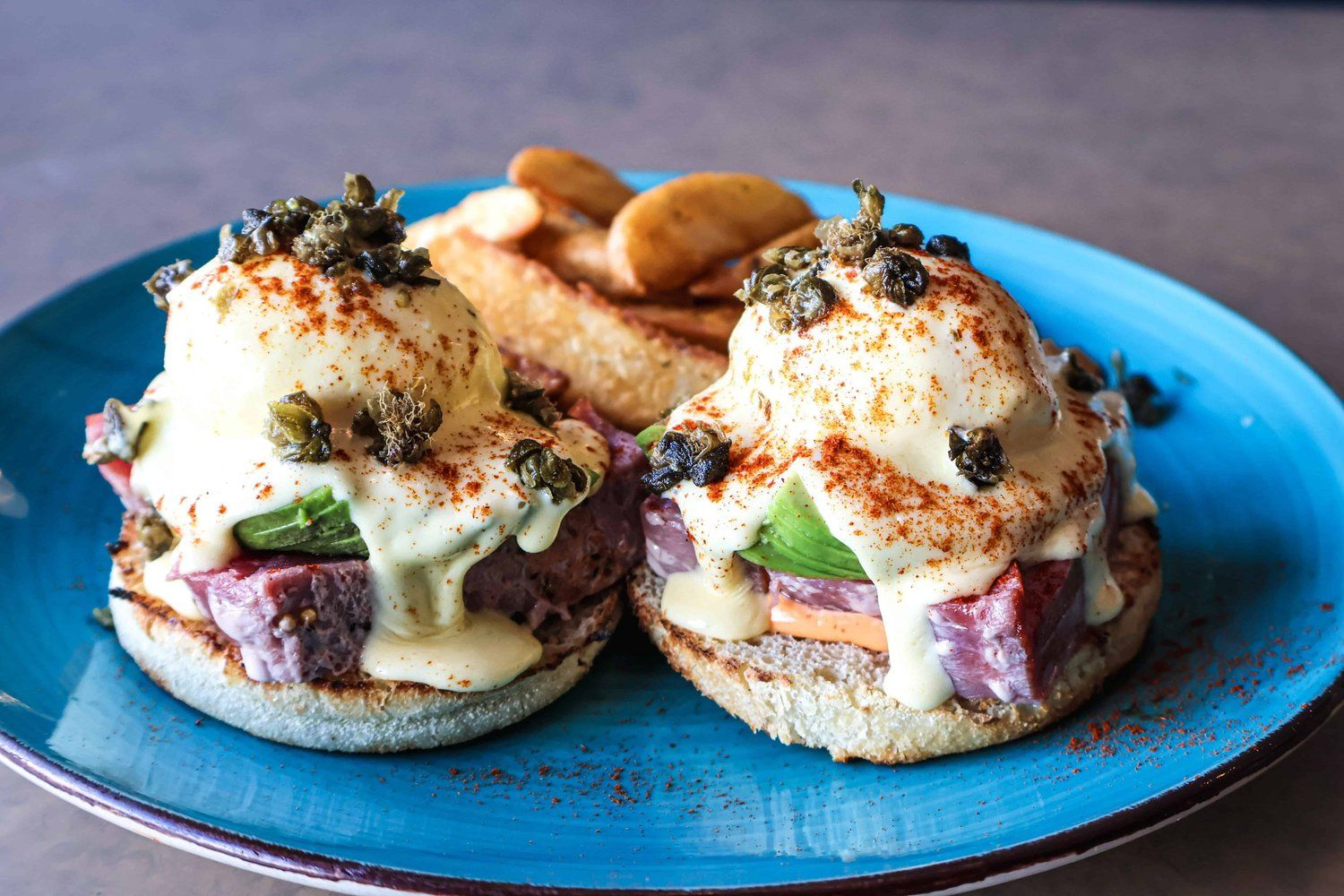 Two gourmet breakfast sandwiches topped with fried eggs and herbs on a blue plate, with potato wedges behind them