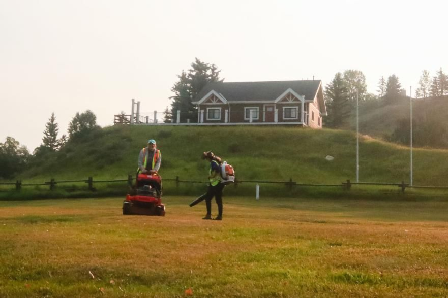 Two workers mowing a grassy field in front of a house on a hill at sunset
