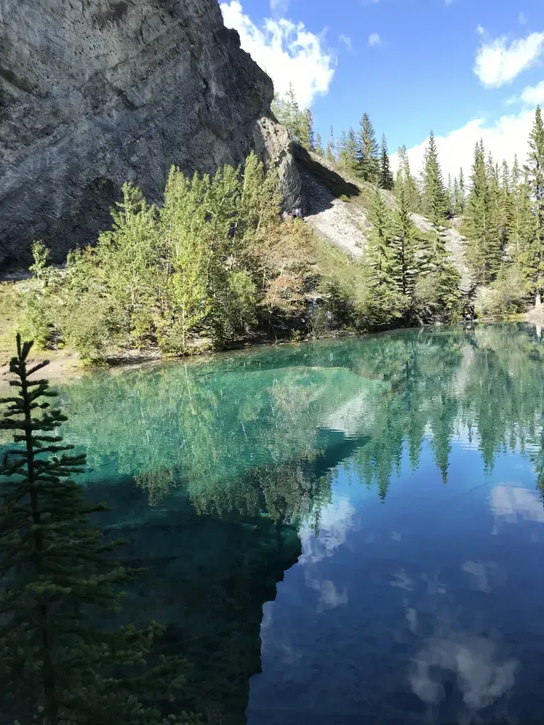 Emerald lake reflecting a rocky cliff and evergreens under a blue sky.