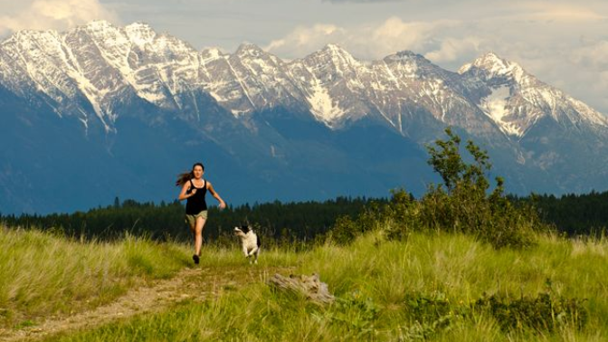 A person jogs with a dog on a grassy trail in front of a scenic backdrop of snow-capped mountains.