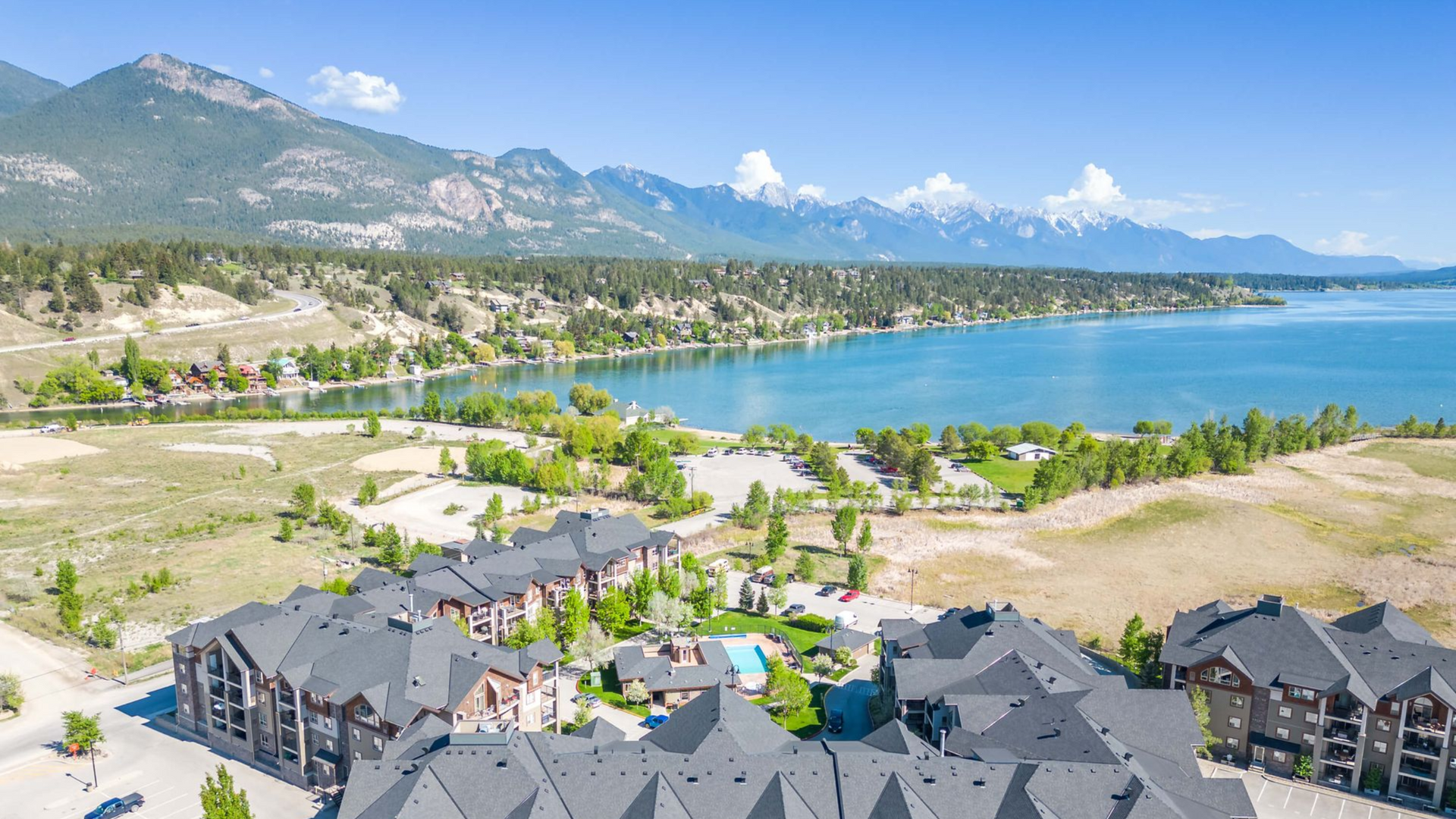 Aerial view of a multi-story lodge nestled near a scenic lake, surrounded by mountains under a clear blue sky.