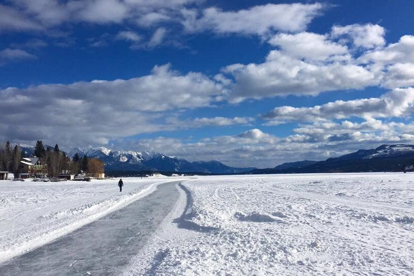 Snow-covered frozen lake with a winding trail, distant mountains, and a person walking under a blue cloudy sky