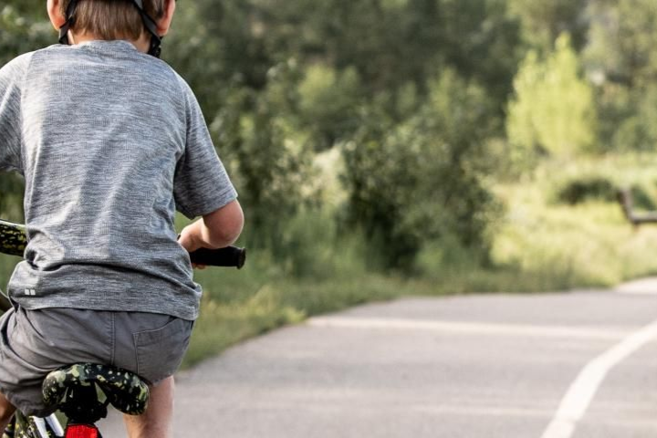 Child riding a bicycle on a paved path beside green trees and grass