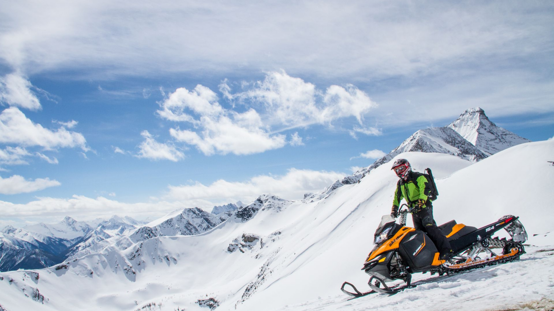 A snowmobiler in a green jacket sits on a yellow snowmobile atop a snowy mountain peak under a partly cloudy sky.
