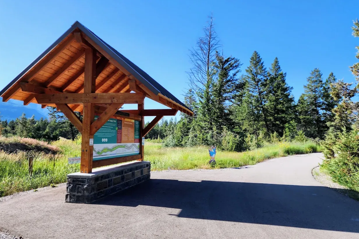 Wooden trail sign shelter beside a paved path, with trees and blue sky in a sunny park.