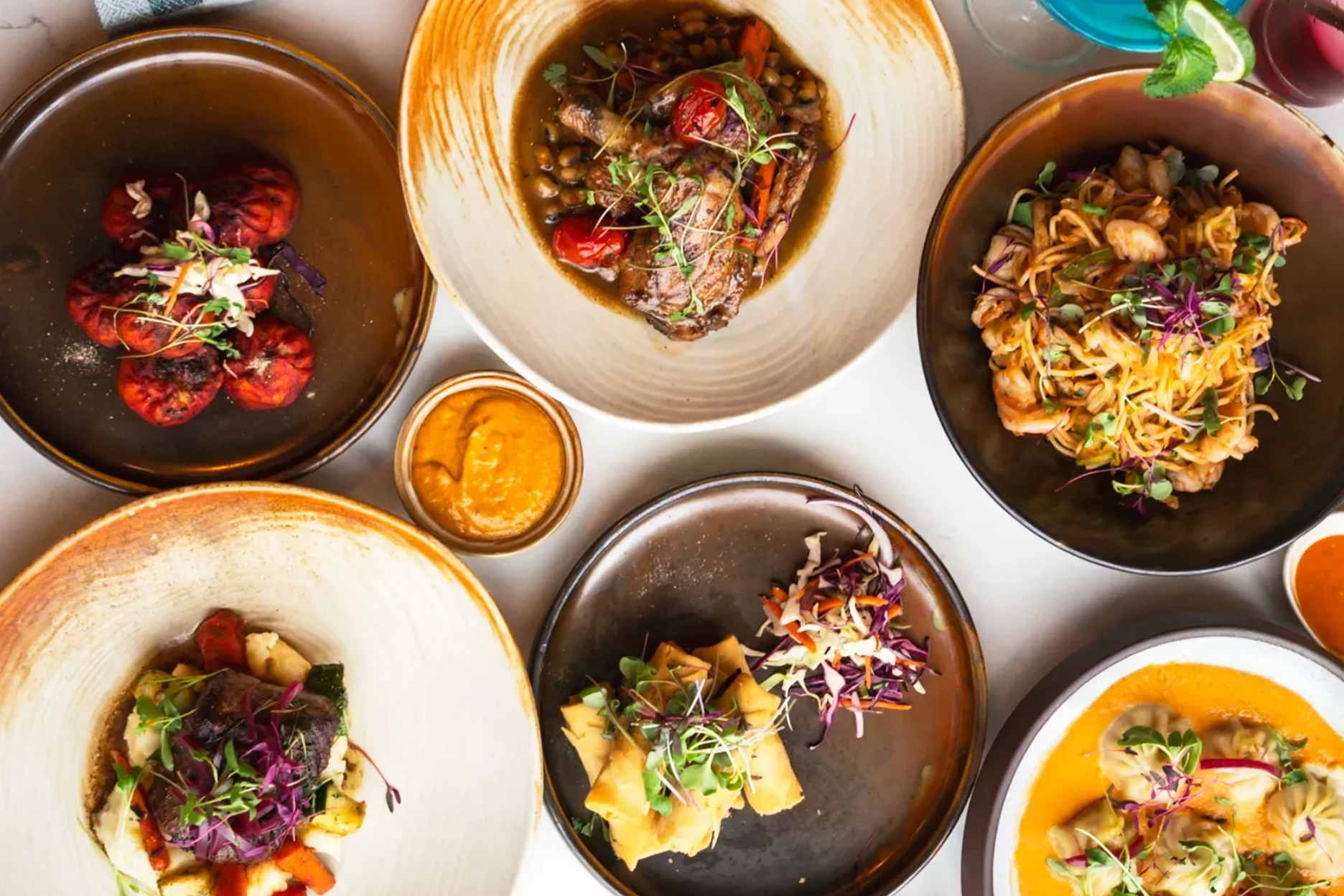 Overhead view of six colorful plated dishes arranged in a circle on a white table