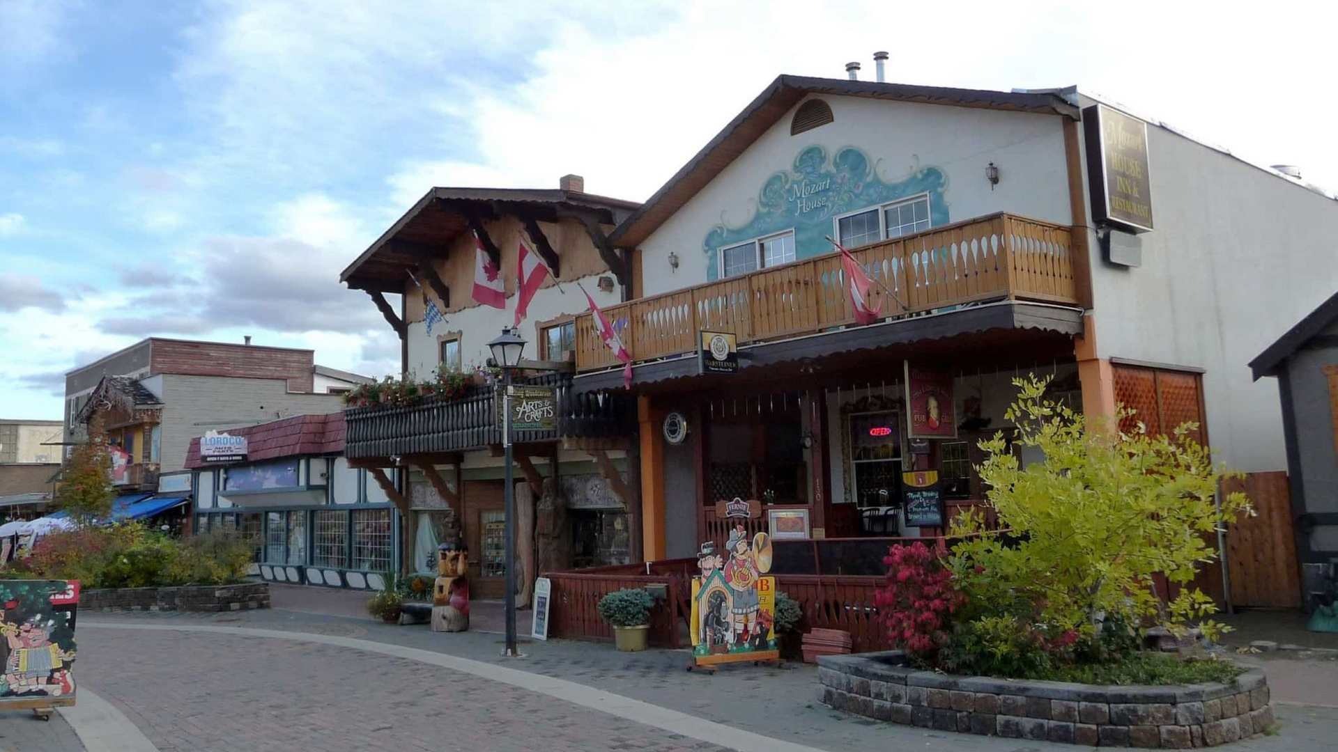 Outdoor market storefronts with colorful signs, a wooden balcony, and a cobblestone walkway.