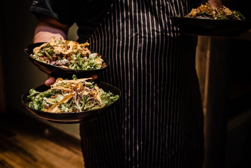 Person in black and white striped apron carries three bowls of food, possibly salads, indoors.