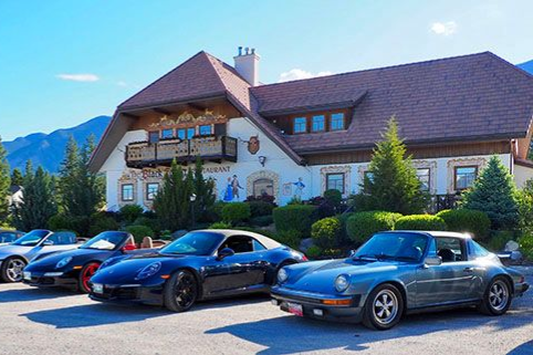 A chalet-style house with several parked sports cars on a sunny mountain roadside