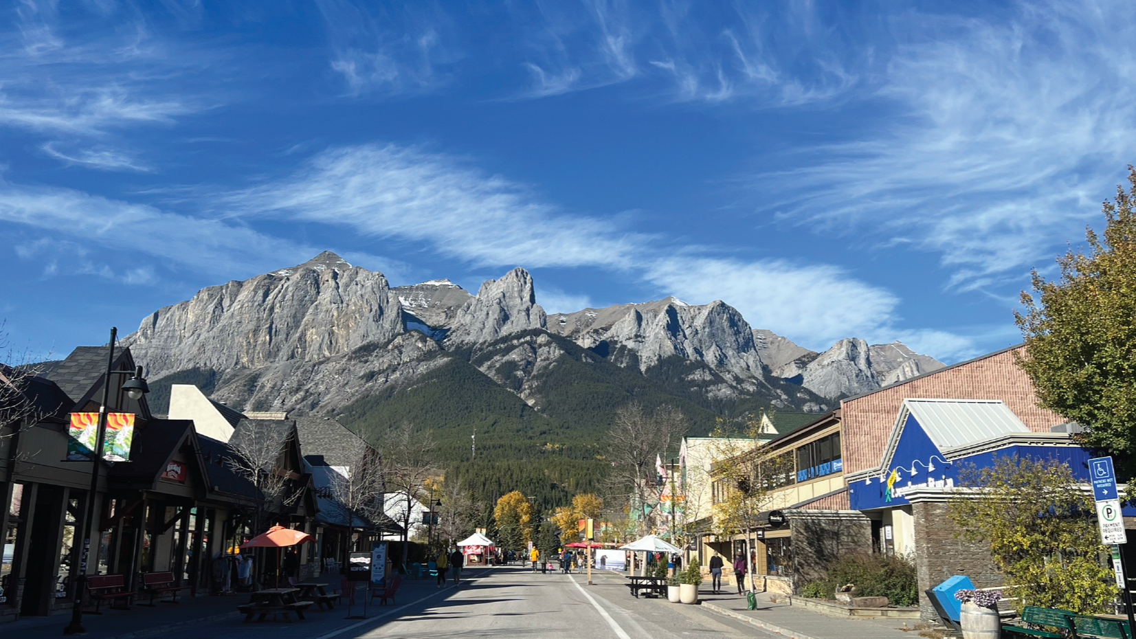 A street view in a mountain town, featuring shops on either side beneath a large, rocky mountain range under a blue sky.