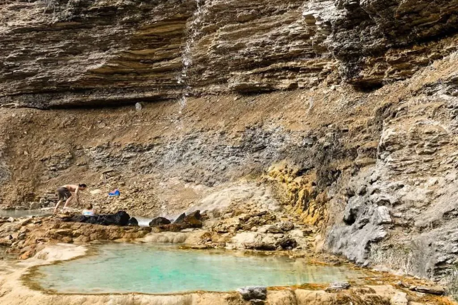 Rocky cliff with a small turquoise pool and a thin waterfall in a cave-like alcove