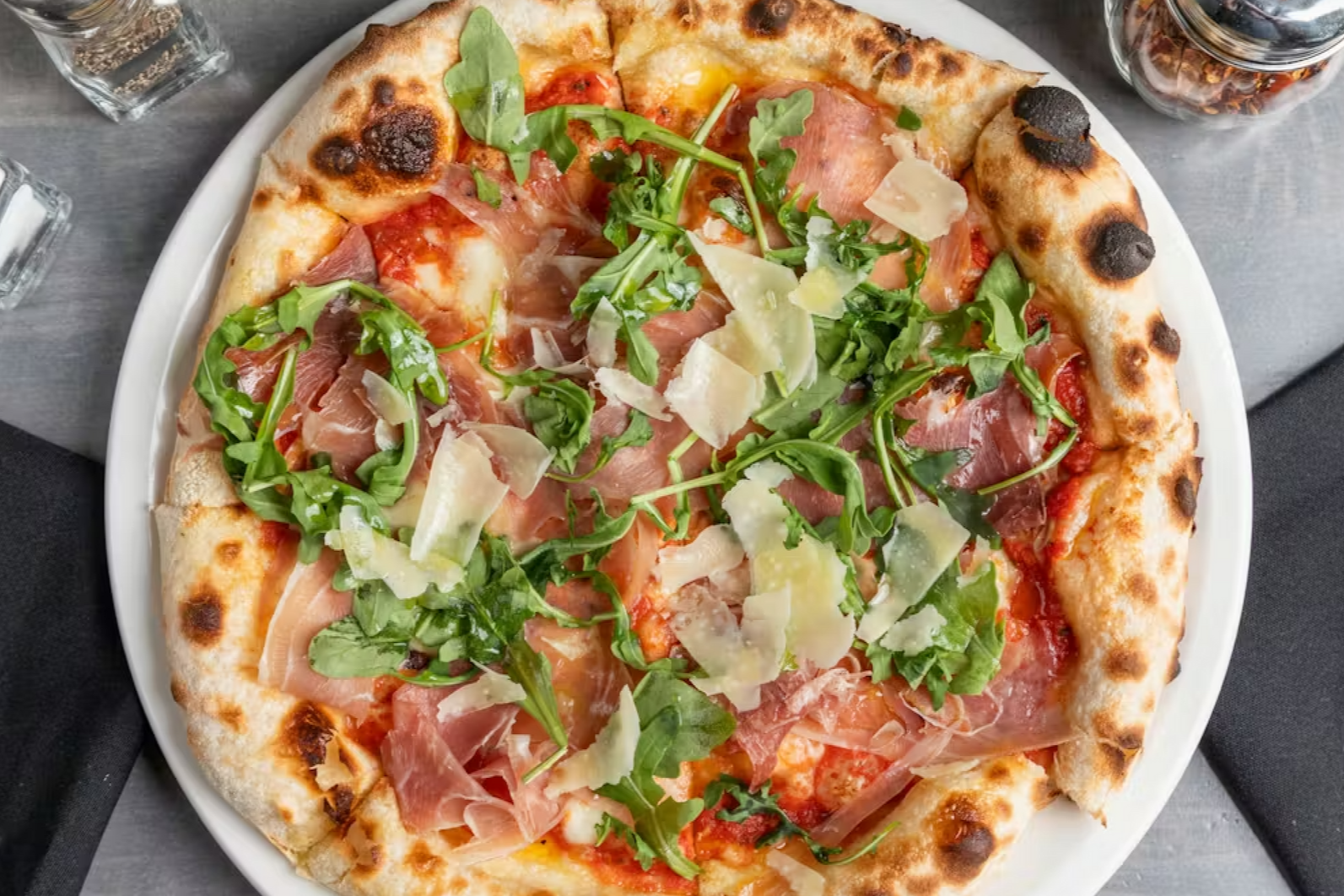 Neapolitan pizza topped with arugula, prosciutto, and shaved cheese on a white plate