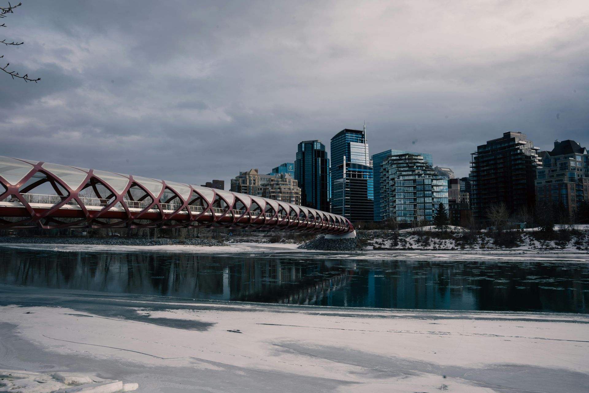 Red-arched bridge over icy river, leading to a cityscape under a cloudy sky.