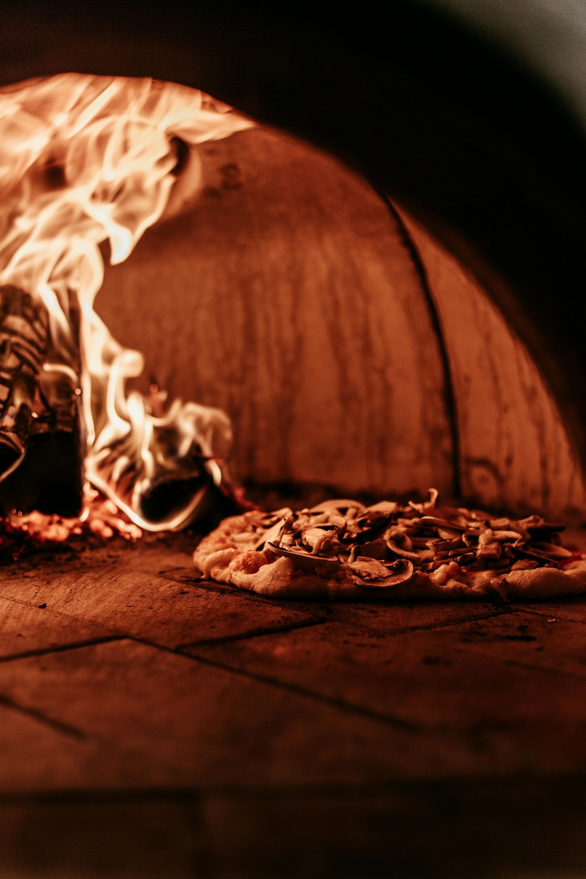 Pizza baking in a wood-fired oven; flames next to the pizza in a dimly lit, brick interior.
