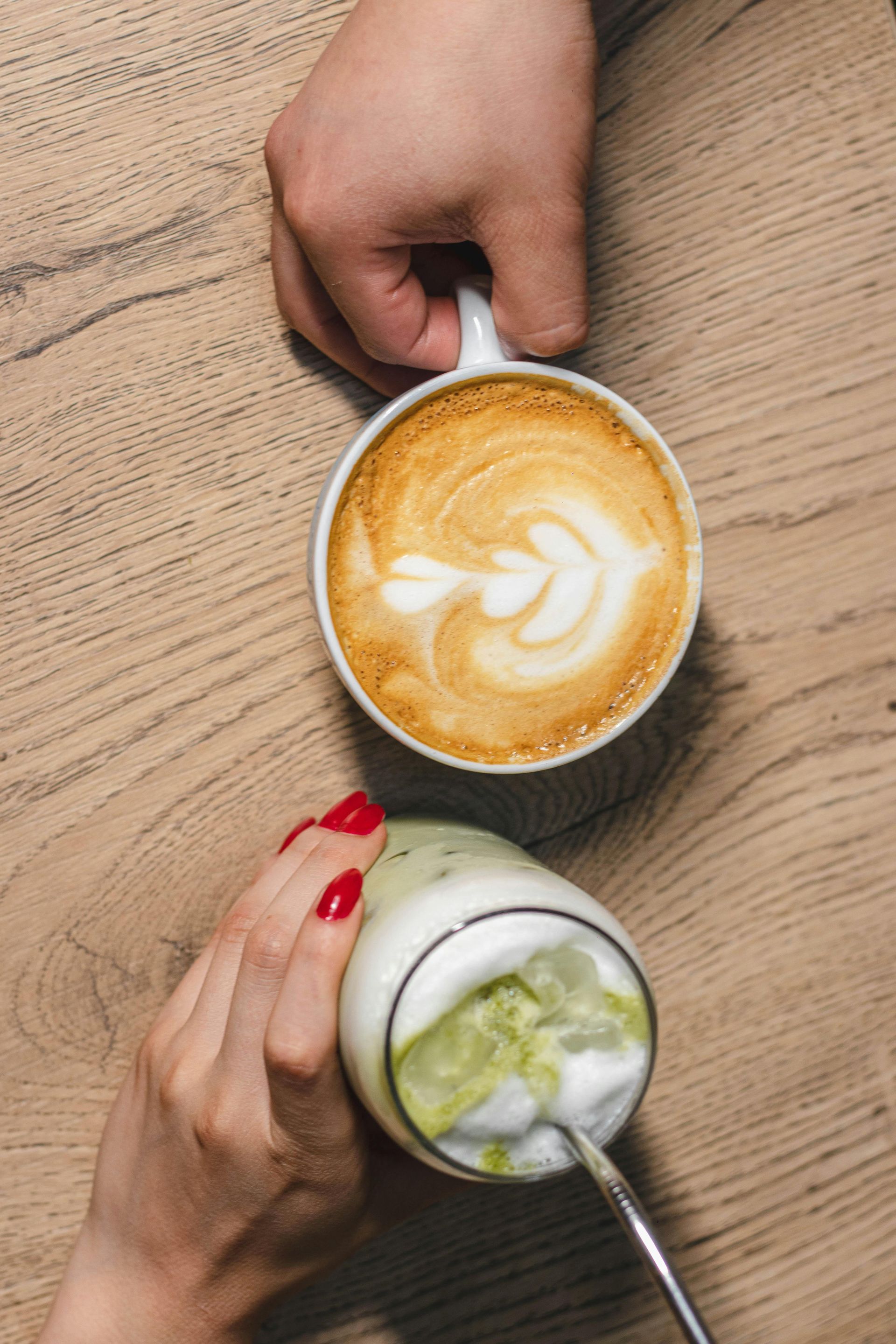 Hands holding a latte with art and a matcha drink with foam; table setting.