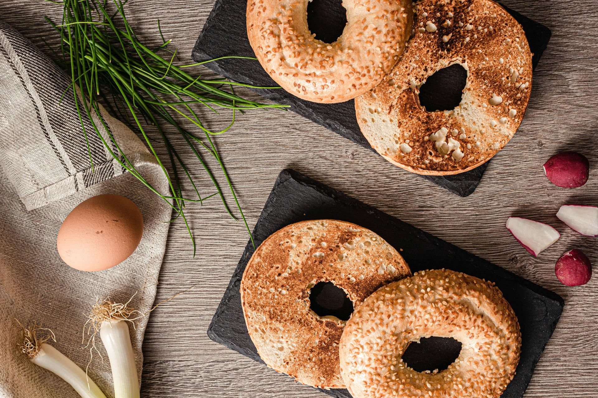 Bagels with sesame seeds on black boards, with chives, radishes, and an egg.