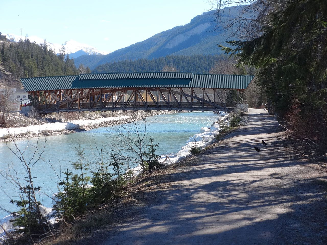 Wooden covered bridge over turquoise river, mountain backdrop. Pathway in foreground.