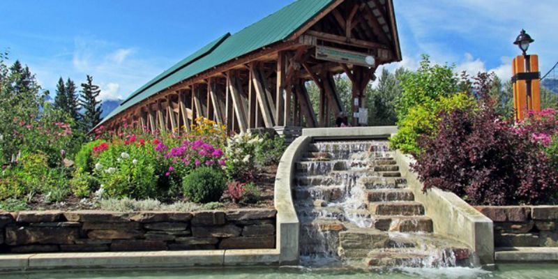 Covered wooden bridge with waterfall in front, surrounded by flowers and lush greenery.