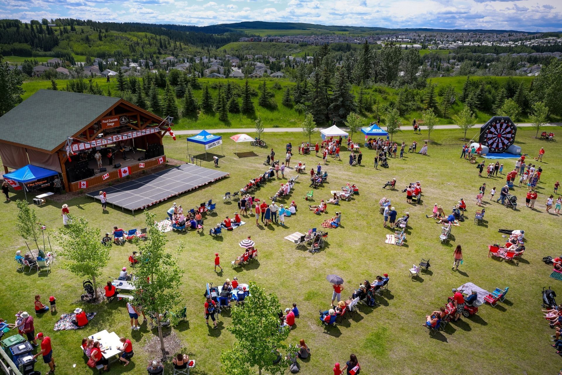 Outdoor festival crowd on a grassy field beside a wooden stage, with tents and trees in the background.