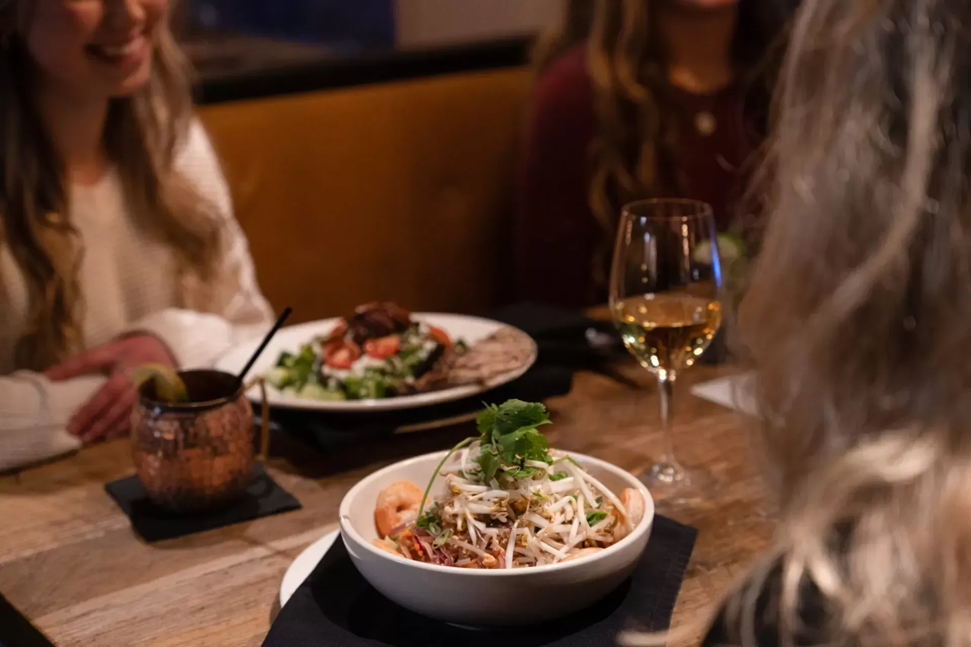 Dining table with bowls of pasta, wine glasses, and two people chatting in a warmly lit restaurant