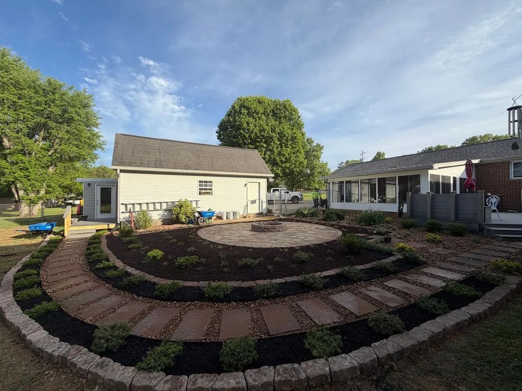Circular brick garden beds in a landscaped yard with a small cream house and patio seating under a blue sky