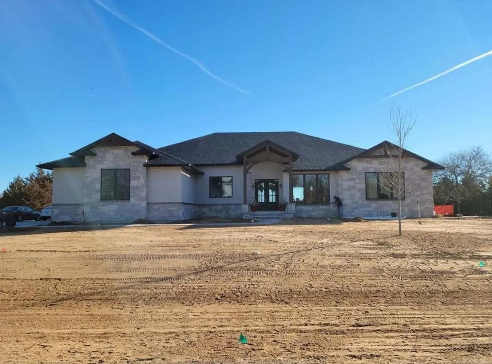 Single-story house under construction with gray stone exterior and a bare dirt front yard under a blue sky