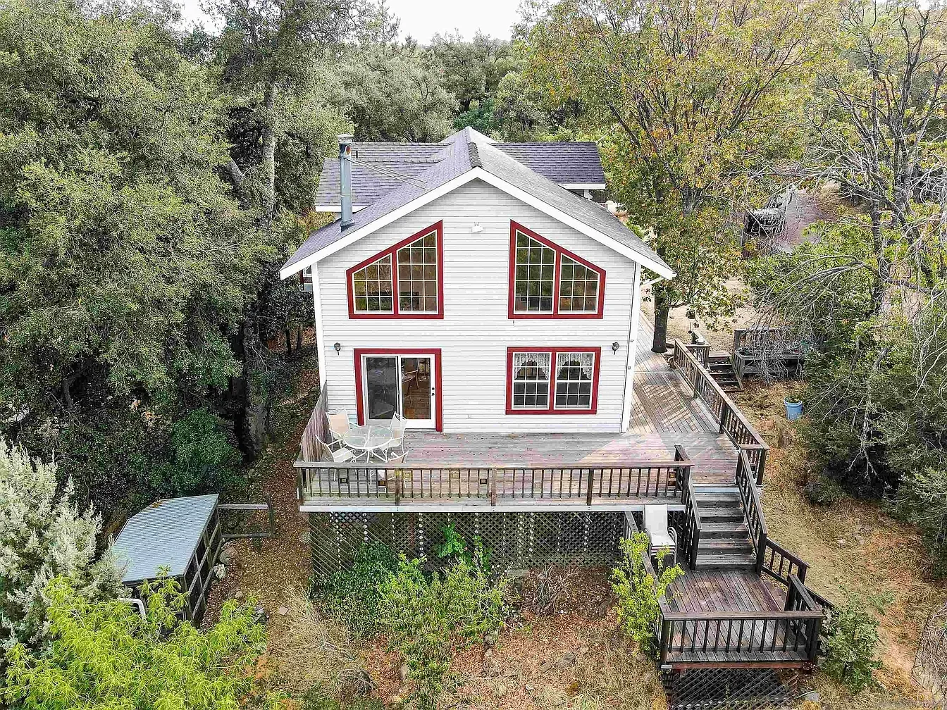 White house with red-trim windows and a deck, surrounded by trees on a wooded hillside.