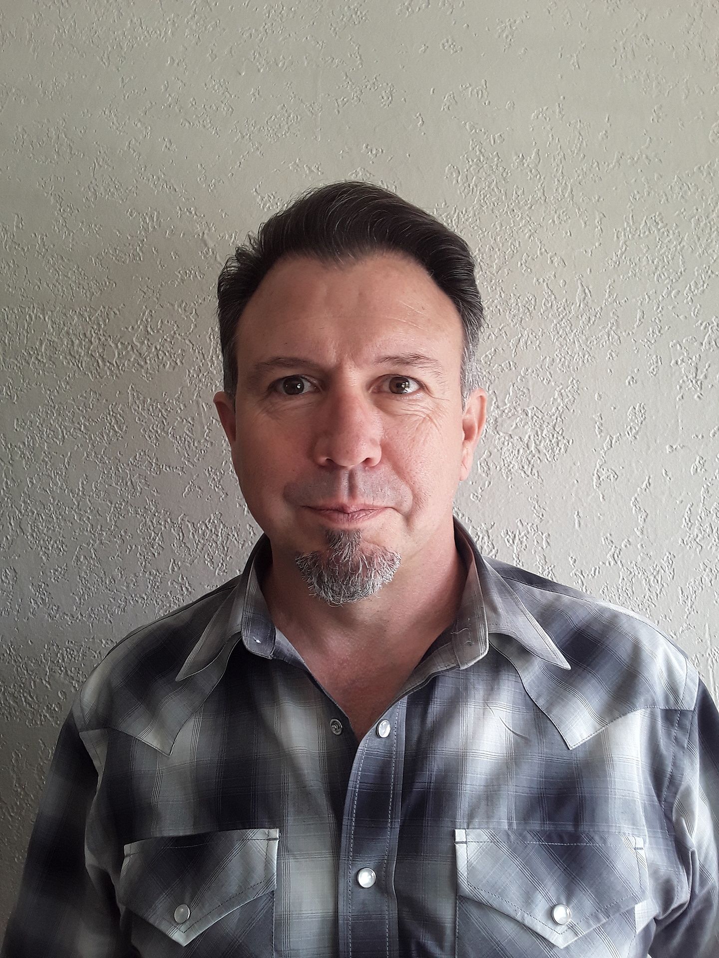 Man standing indoors against a textured wall, wearing a gray plaid shirt, and looking at the camera.