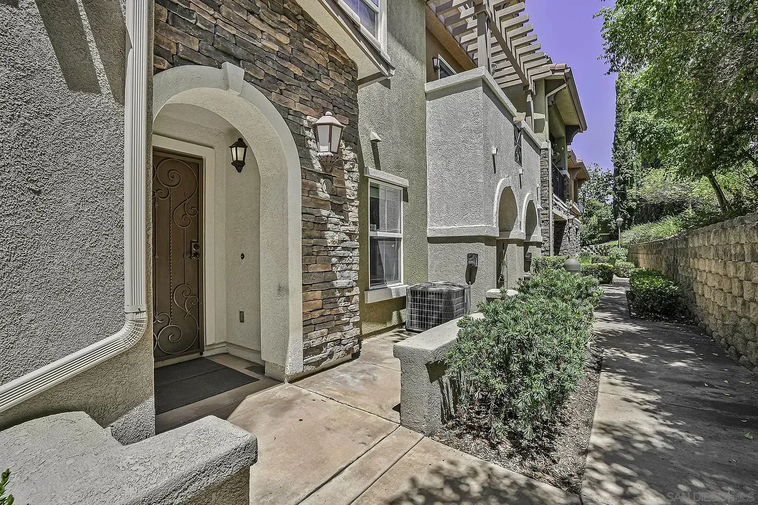 Stone house entrance with arched doorway, narrow path, and shrubs beside a textured wall