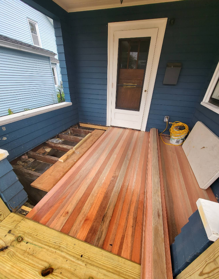 Porch with new wooden flooring being installed, blue house exterior in background.