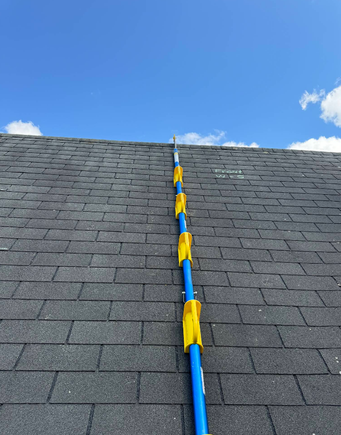 Blue pole with yellow clips extends from a dark shingled roof towards a blue sky with clouds.