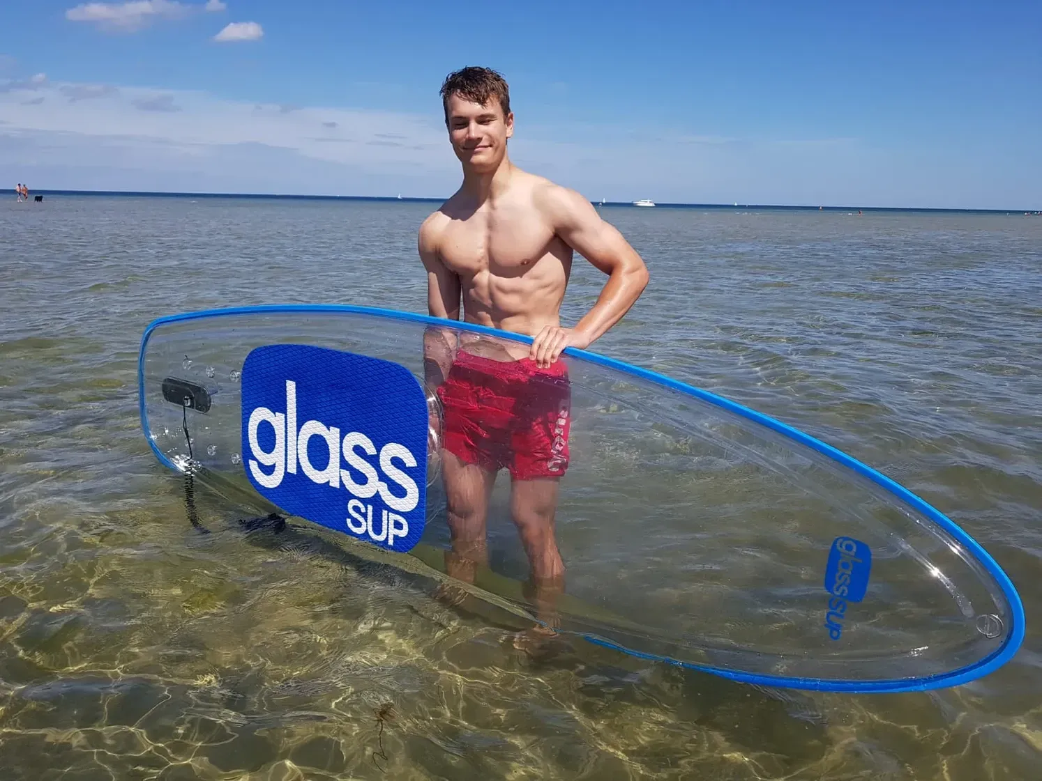 Ein sehr muskulöser  Mann  steht im Wasser und hält ein durchsichtiges glassSUP.