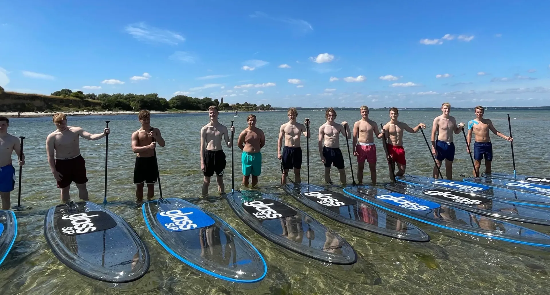Eine Gruppe Männer stehen aufgereiht vor den glassSUPs im glasklaren Wasser.