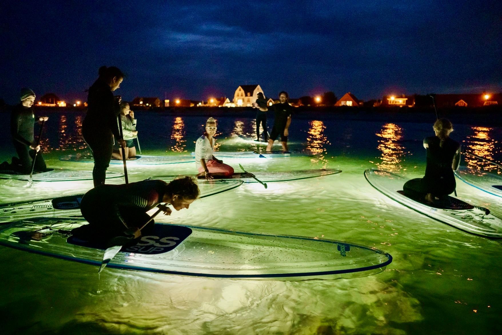 Eine Gruppe von Menschen steht nachts auf transparenten Paddelbrettern im Wasser und schaut in die Unterwasser Welt.
