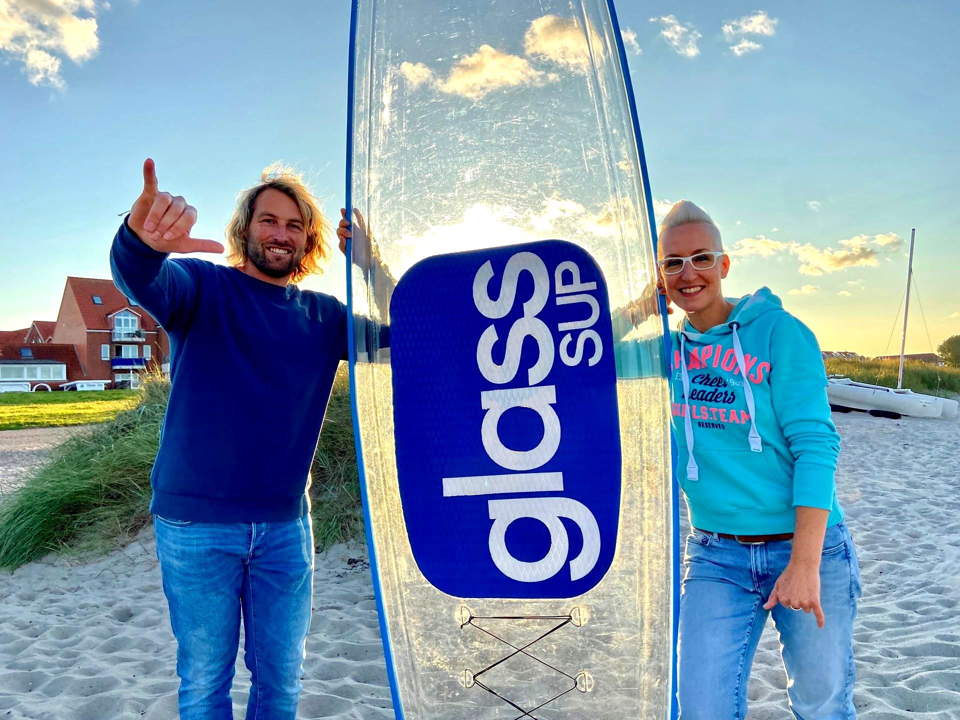 Ein Mann und eine Frau stehen an einem Strand und halten ein gläsernes SUP-Surfbrett.