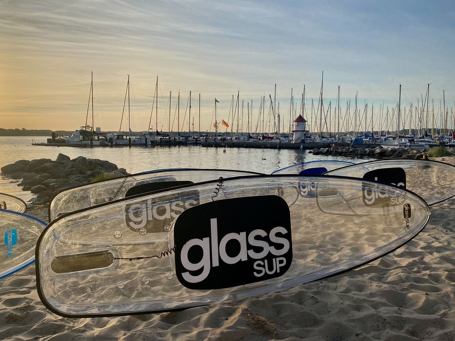 Zwei glassSUP-Boards liegen auf einem Sandstrand und im Hintergrund  ein Hafen mit Segelschiffen und Segelmasten